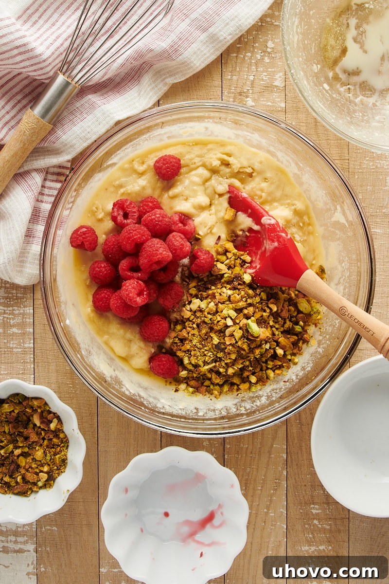 Overhead view of muffin batter with raspberries and pistachios freshly added, showing the colorful inclusions beginning to be folded in.