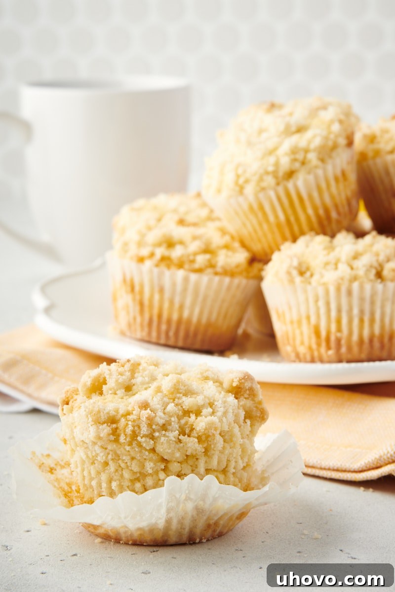 A perfectly baked Lemon Crumb Muffin, unwrapped and bitten, with more muffins artfully arranged on a plate in the background.