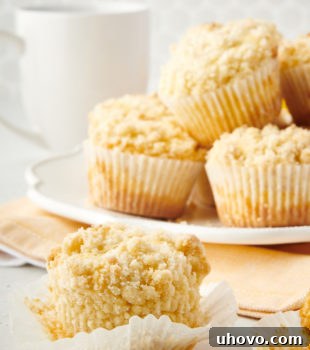 An unwrapped lemon crumb muffin with a plate of muffins in the background, highlighting the soft interior and crumbly top.