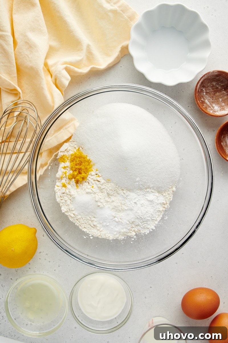 Overhead view of dry ingredients including flour, sugar, baking powder, baking soda, salt, and lemon zest in a glass mixing bowl.