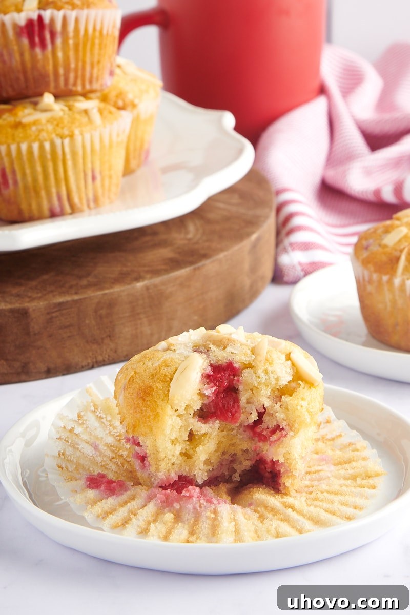 A sliced Red Currant Muffin on a white plate, showing the berries inside, with more muffins in the background