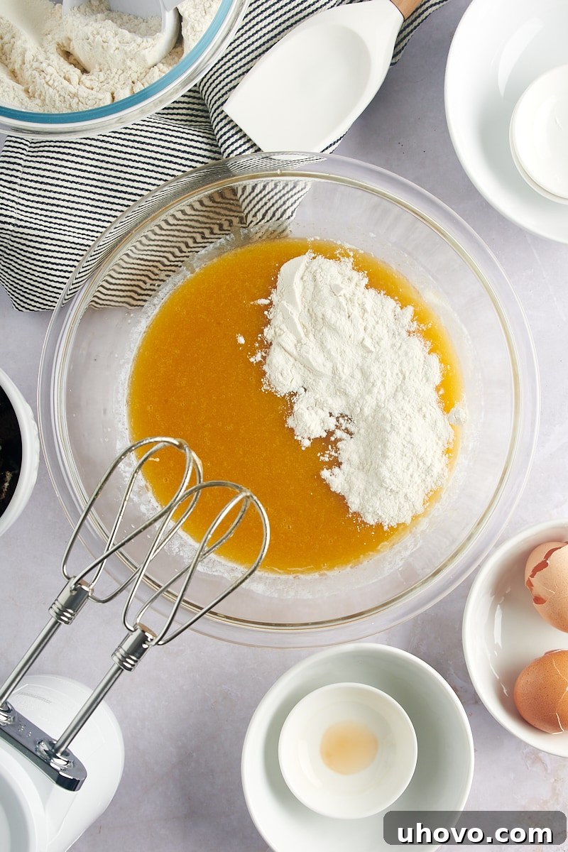 Overhead view of flour being added gradually to the wet blondie batter in a glass mixing bowl.
