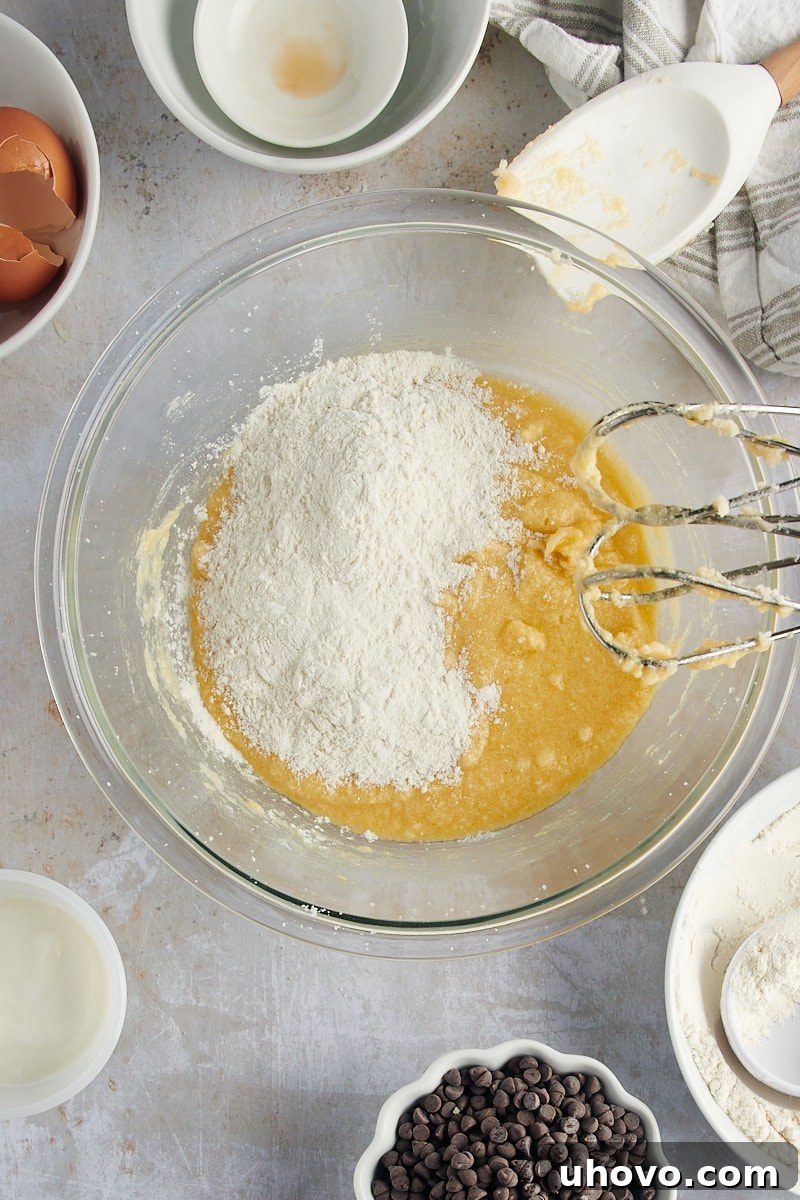 An overhead view shows the dry ingredients being gradually added to the wet mixture in a glass mixing bowl, illustrating the careful integration of components for Mini Chocolate Chip Bundt Cakes.