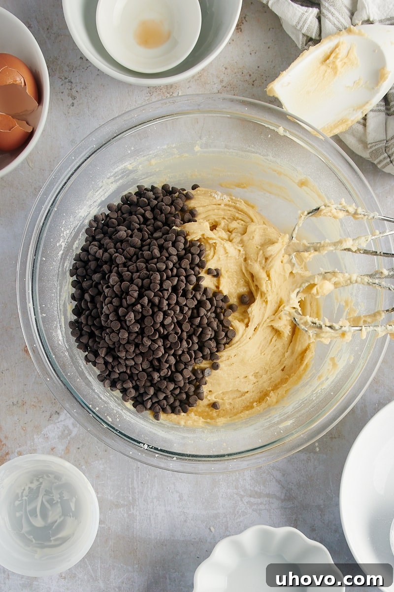 An overhead view of mini chocolate chips being gently folded into the rich cake batter in a glass mixing bowl, preparing the Mini Chocolate Chip Bundt Cakes for baking.