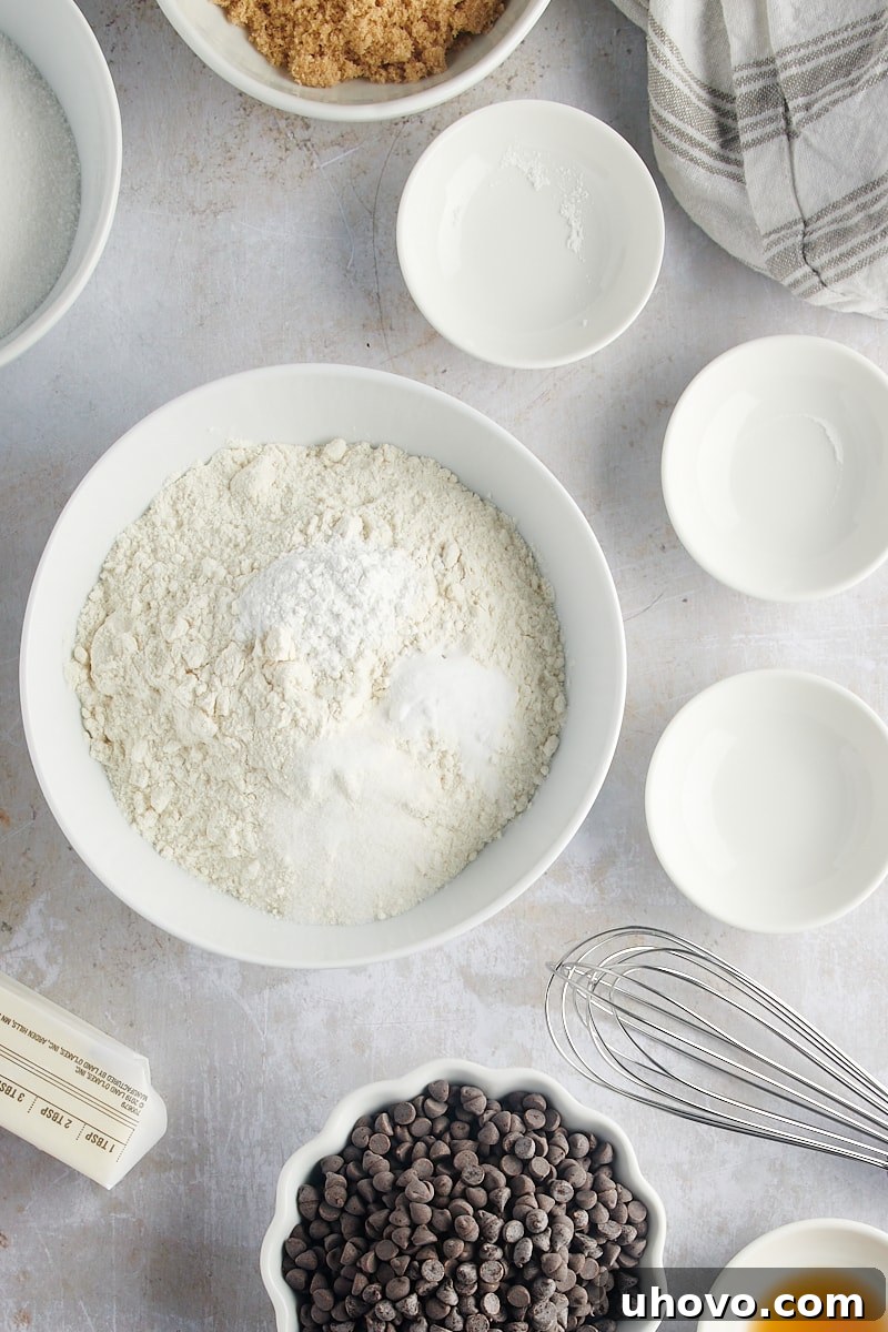 An overhead view of flour, baking powder, baking soda, and salt meticulously arranged in a white mixing bowl before being combined, emphasizing the initial step of dry ingredient preparation.
