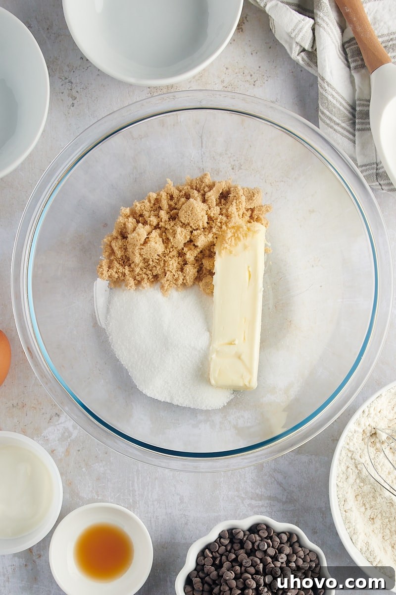 An overhead view of butter, granulated sugar, and light brown sugar being creamed together in a clear glass mixing bowl, highlighting the initial stage of wet ingredient preparation.