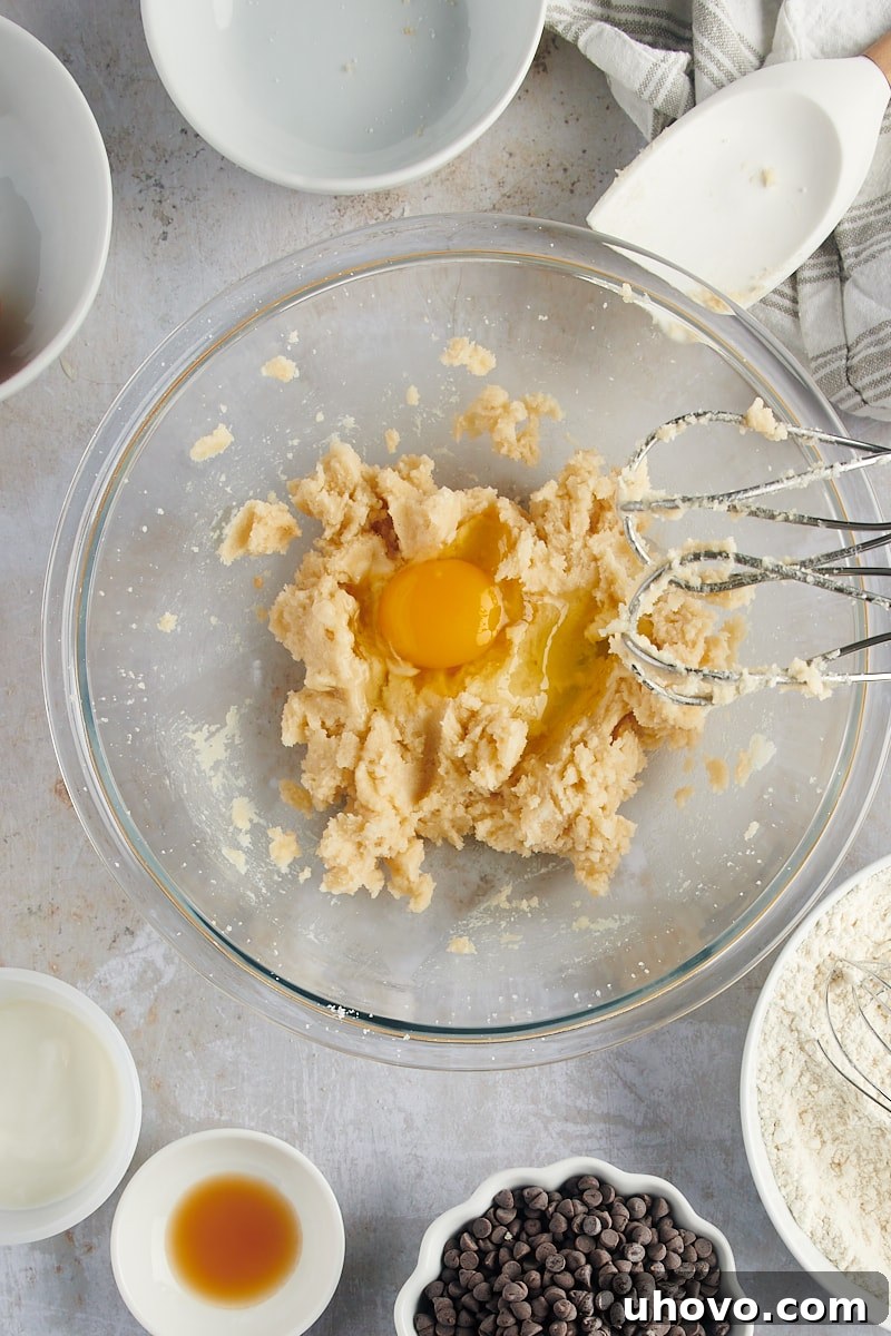 An overhead view captures the moment an egg is carefully added to the creamed wet ingredients in a glass mixing bowl for Mini Chocolate Chip Bundt Cakes, illustrating the step-by-step process.