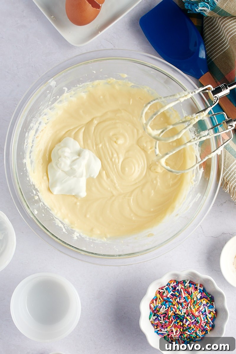 An overhead shot of sour cream being carefully added to the creamy cheesecake batter in a glass mixing bowl, ready for its final incorporation.
