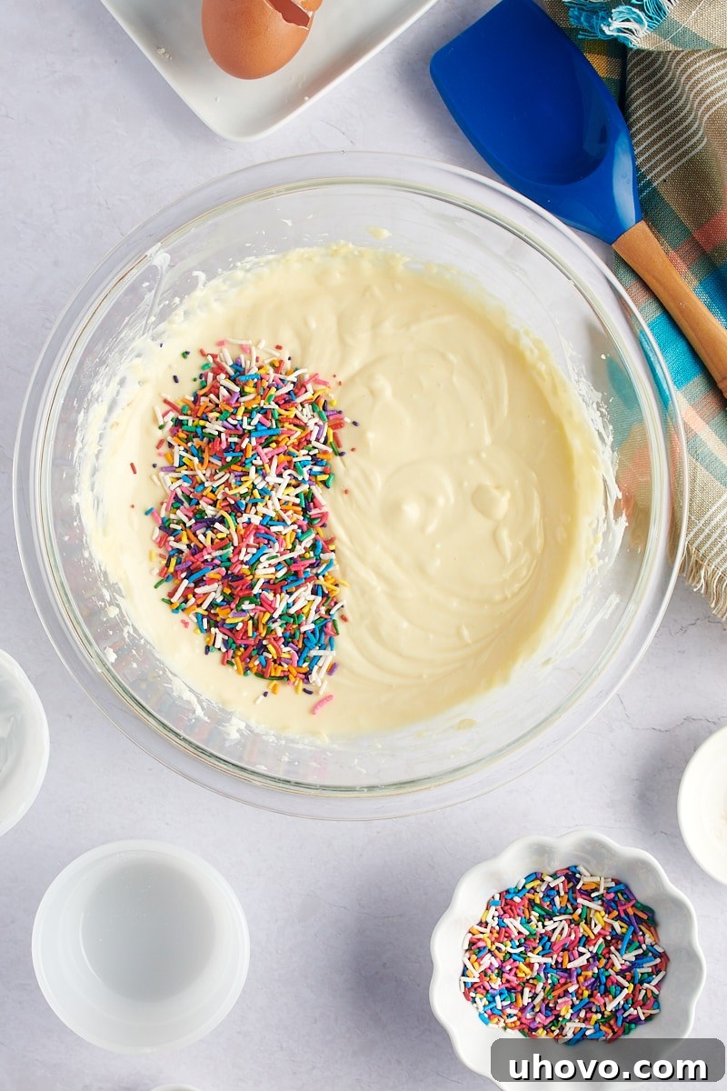 An overhead view of vibrant sprinkles being gently added to the creamy cheesecake batter in a clear glass mixing bowl, preparing for the 'funfetti' effect.