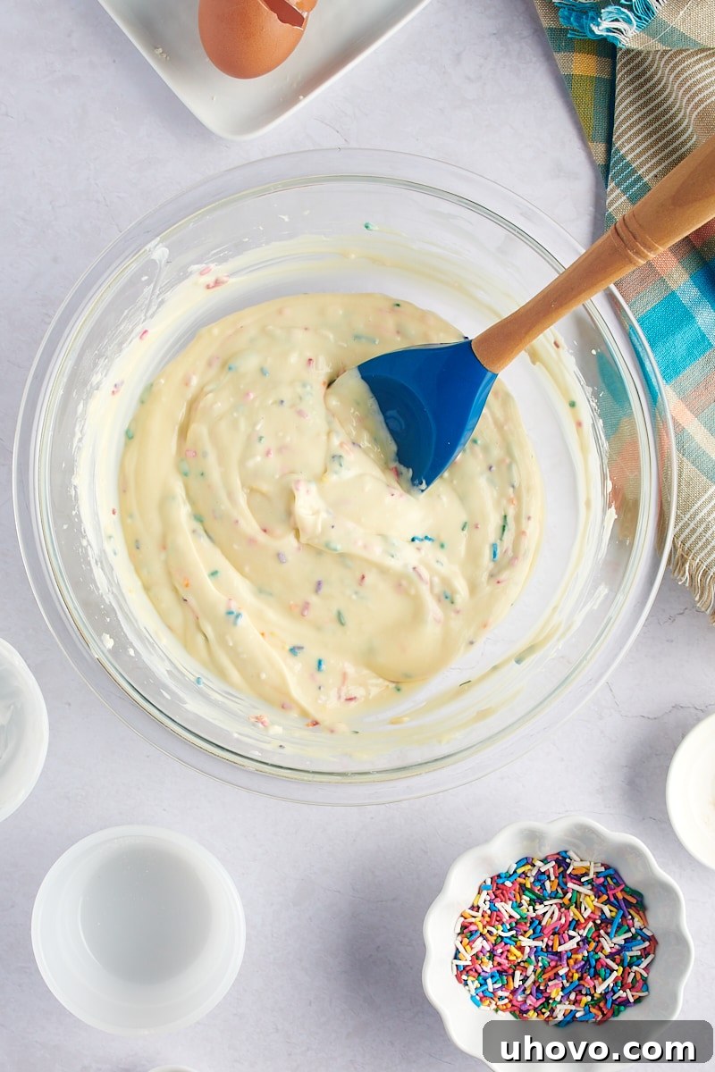 An overhead shot of the thoroughly mixed Mini Funfetti Cheesecake batter, now studded with colorful sprinkles, resting in a glass mixing bowl.