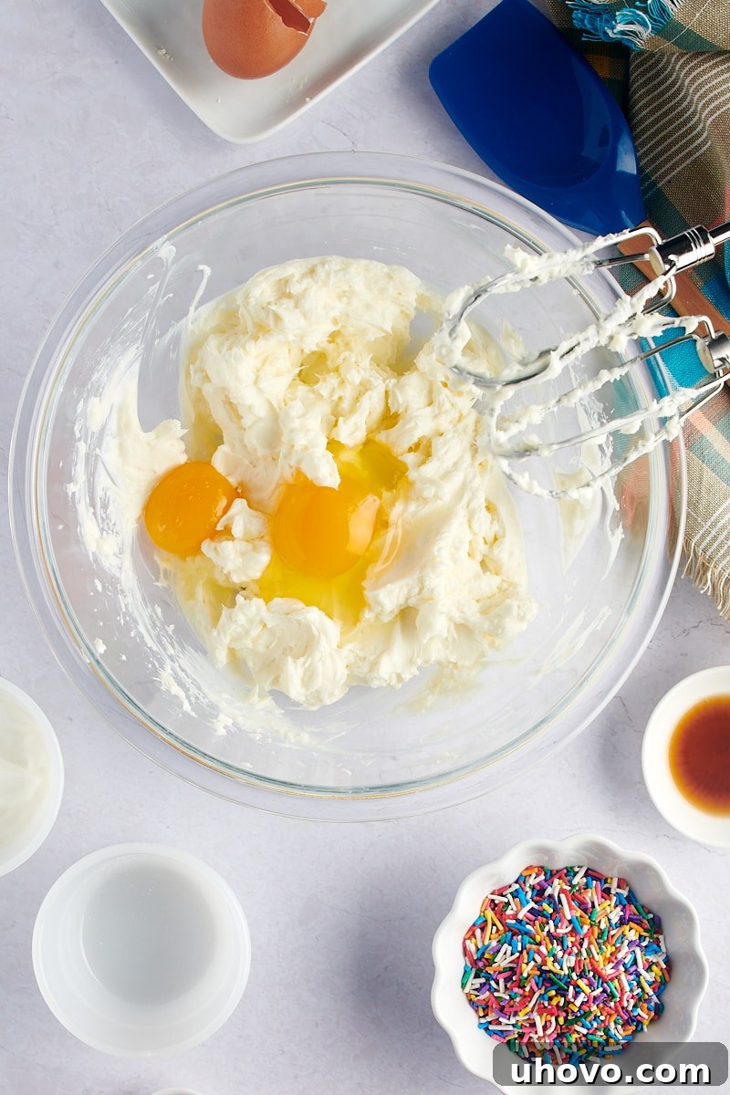 An overhead shot showing eggs carefully cracked into the smooth cheesecake batter within a glass mixing bowl, ready to be incorporated.