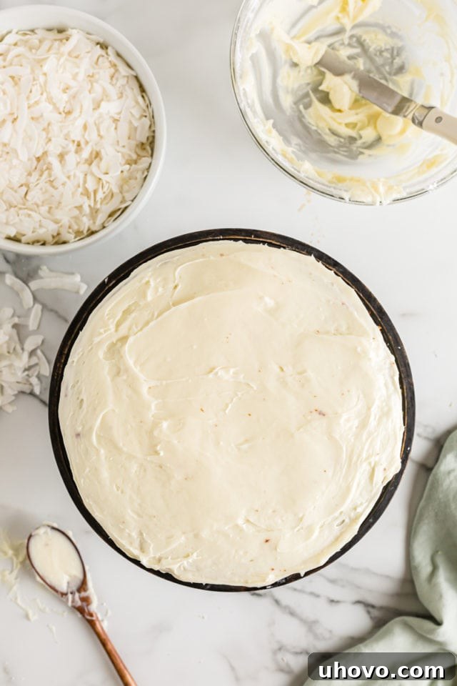 Overhead view of a two-layer coconut cake fully frosted with smooth cream cheese frosting, before the final addition of shredded coconut.