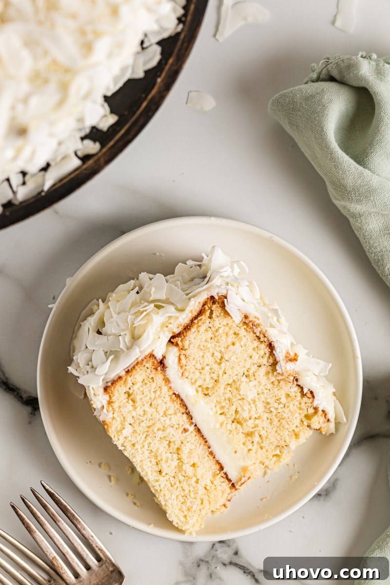A stunning close-up of a single slice of coconut cake on a white plate, with the rest of the beautifully frosted cake visible in the soft-focus background, ready to be enjoyed.