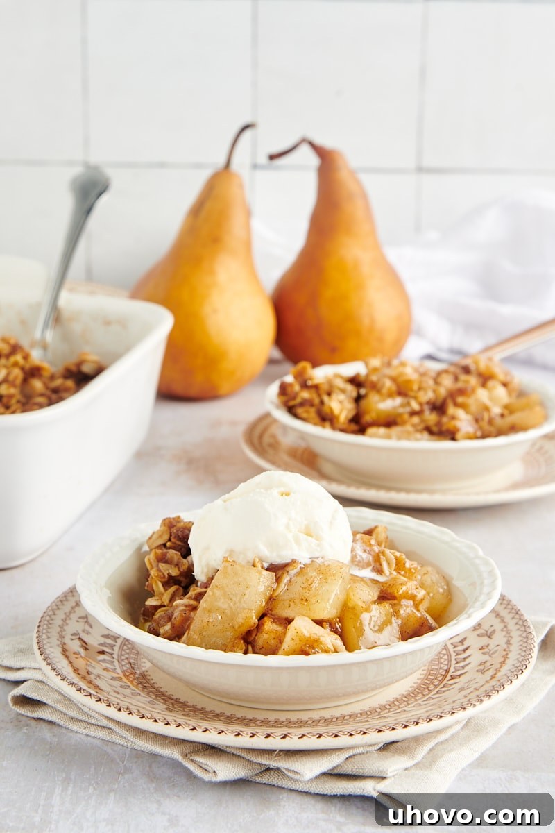 A close-up of Brown Butter Pear Crisp served with a scoop of ice cream in a white bowl, with the baking dish and fresh pears in the soft background.