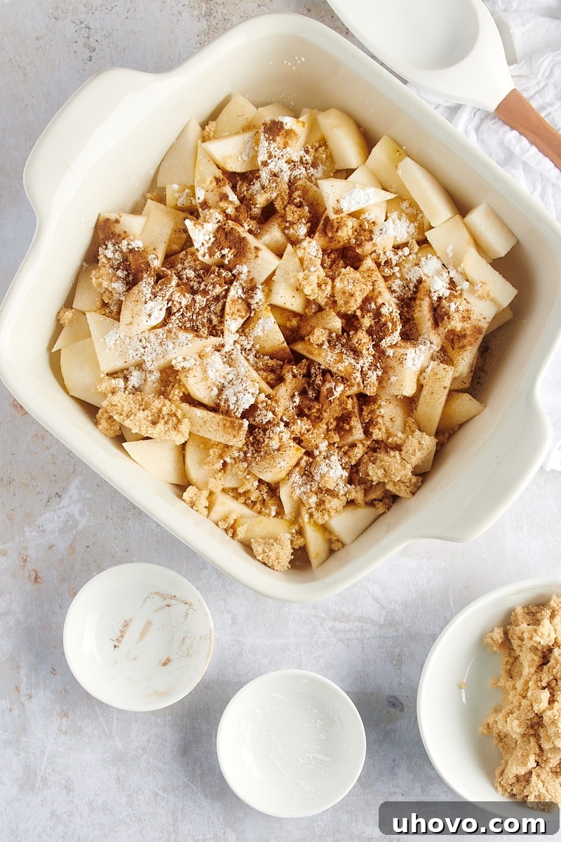 An overhead view captures the start of the filling: neatly sliced pears, golden brown sugar, white cornstarch, and aromatic cinnamon, all waiting to be combined in a white baking dish.
