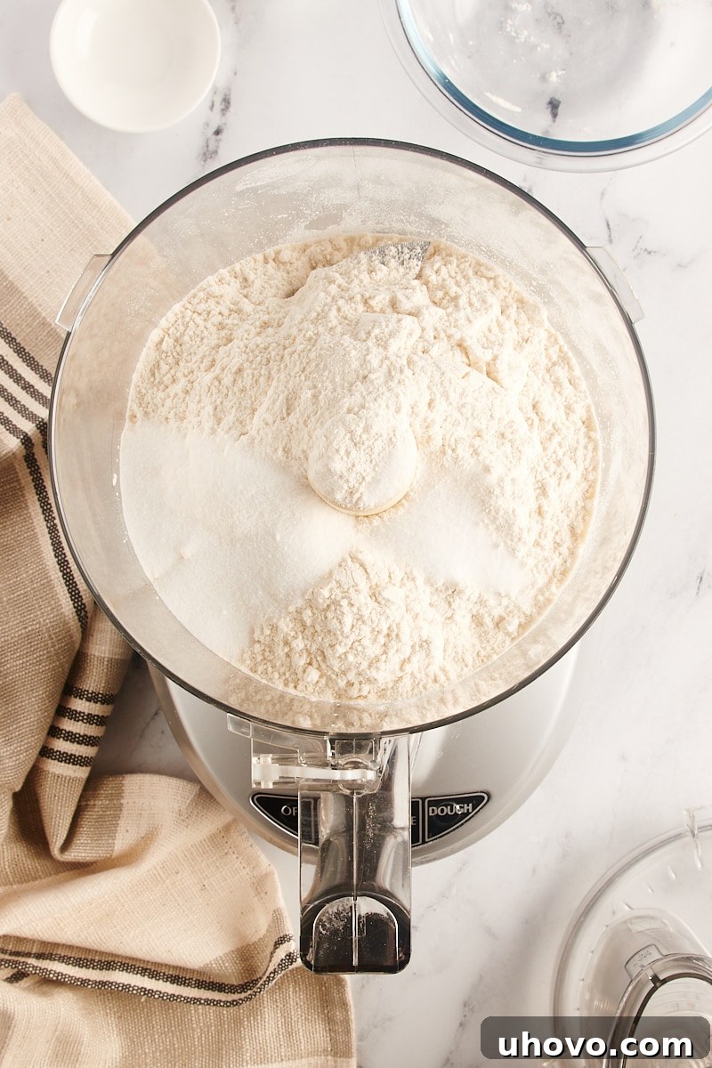 Overhead view of flour, sugar, and salt placed in the bowl of a food processor, ready to be pulsed and combined for the pie crust.