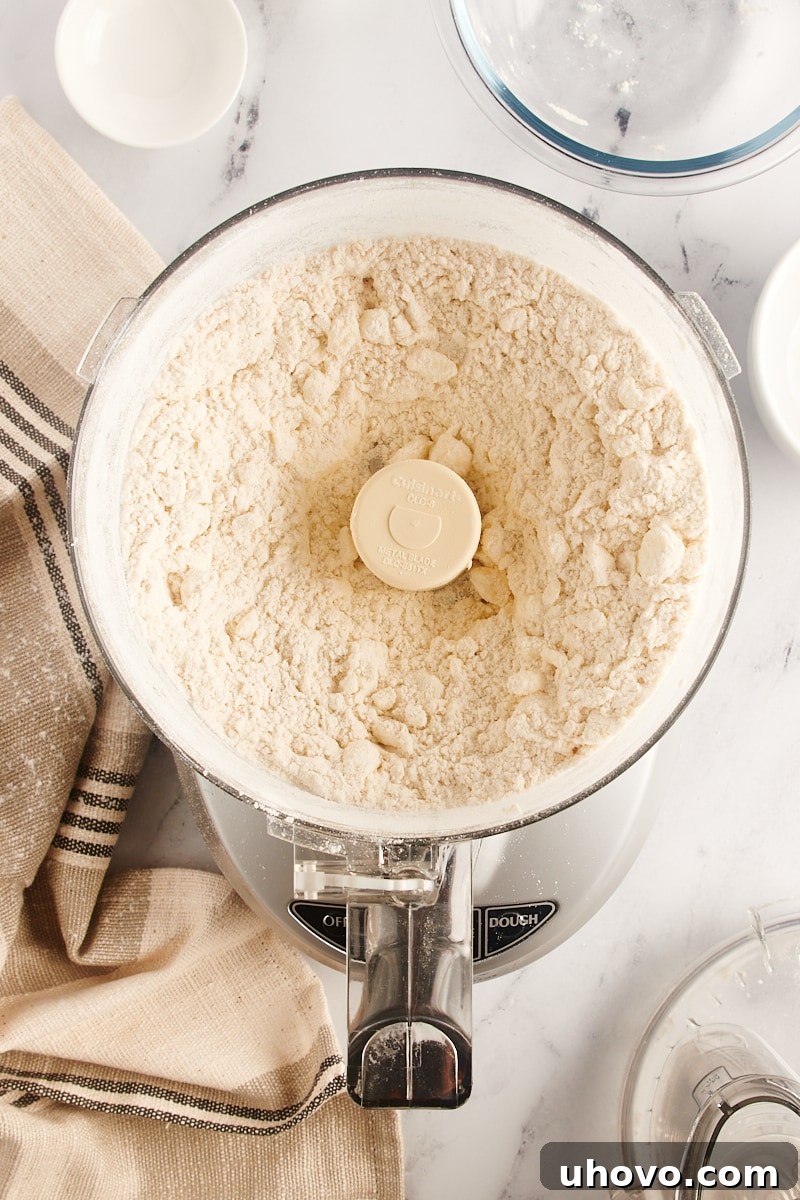 Overhead view of pie crust dough in a food processor after initial pulsing, showing butter pieces that are about the size of large peas, ready for water addition.