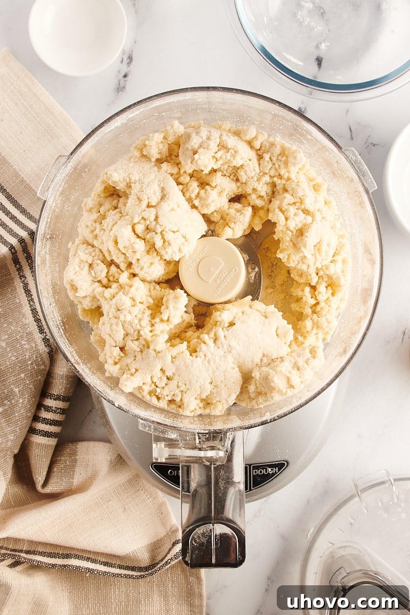 Overhead view of pie crust dough forming large clumps in the bowl of a food processor, indicating it has reached the ideal consistency after adding cold water.