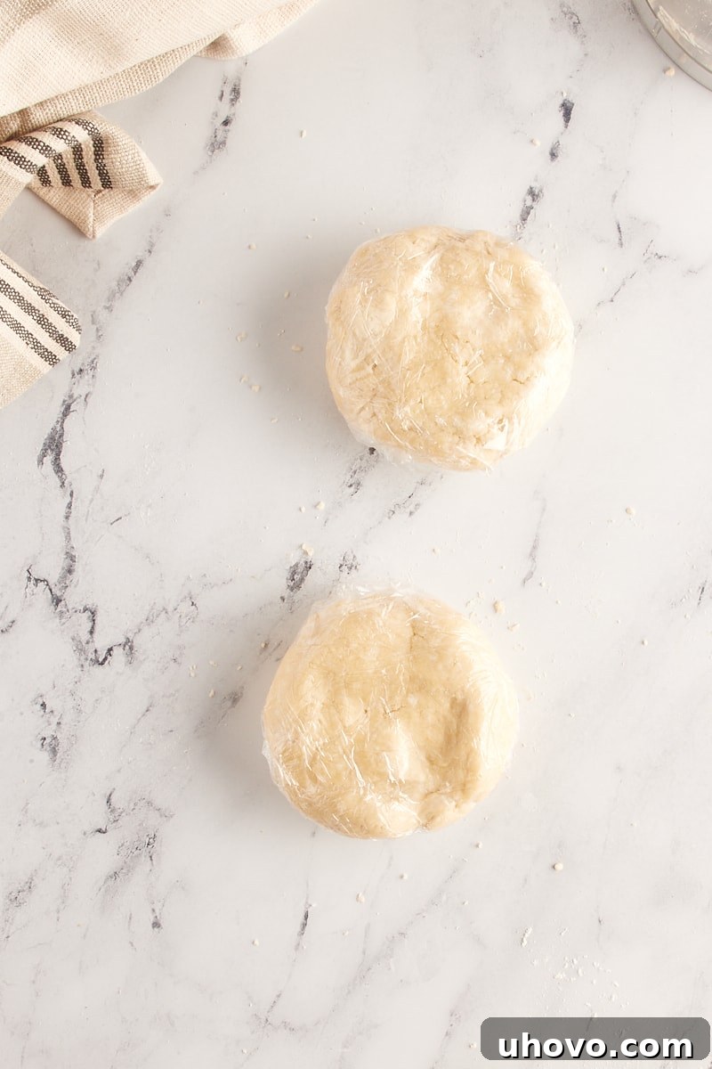 Overhead view of two discs of homemade pie crust dough, flattened and tightly wrapped in plastic wrap, ready to be chilled for optimal texture and ease of rolling.