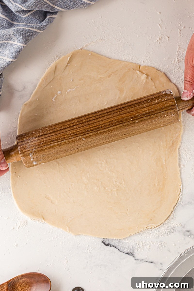 Overhead view of a wooden rolling pin gently rolling out pie crust dough on a floured surface, creating an expanding, even circle.