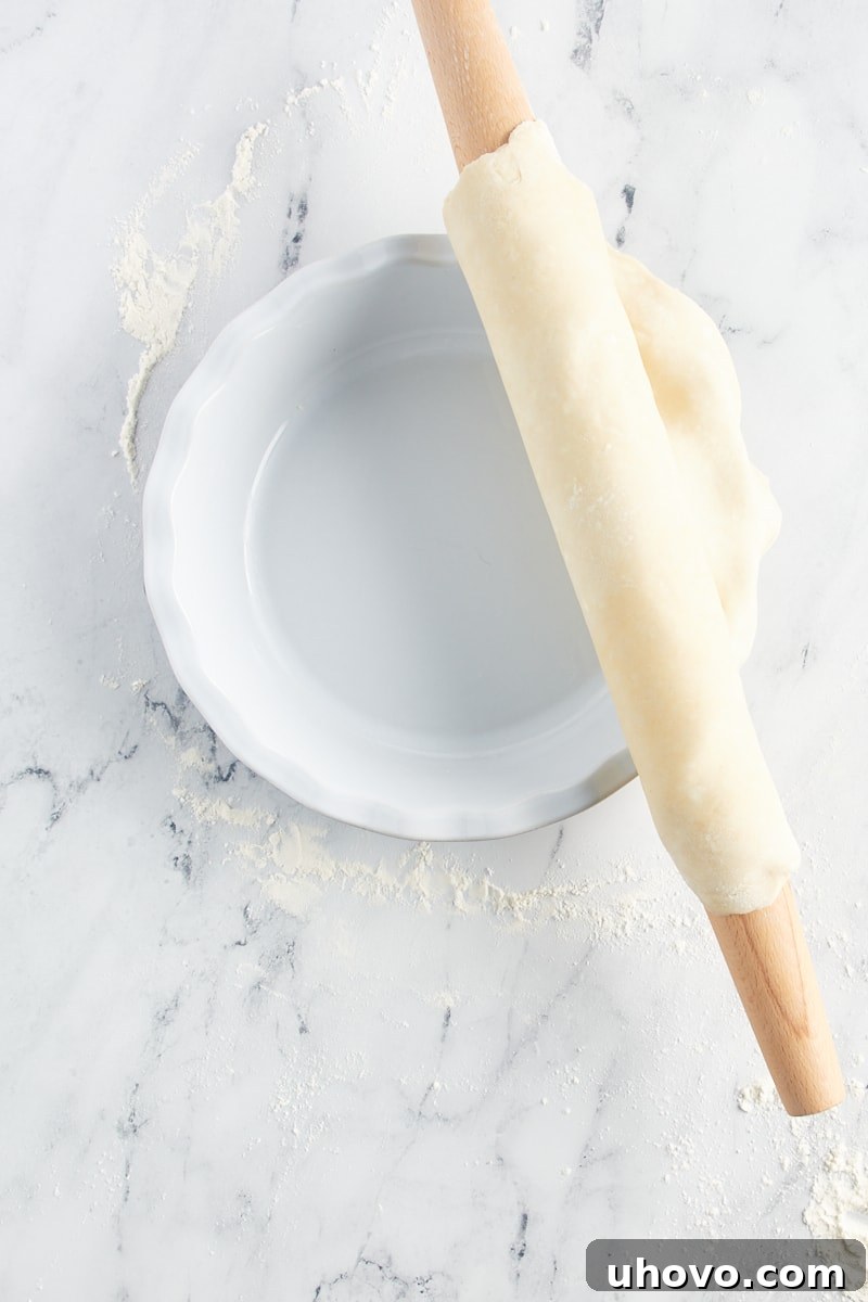 Overhead view of a pie crust rolled onto a rolling pin and being carefully unrolled over a pie plate, demonstrating the transfer technique.