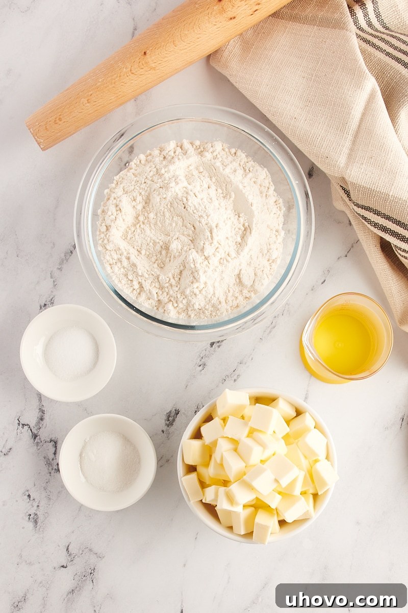 Overhead view of pie crust ingredients: all-purpose flour, granulated sugar, salt, cubed cold unsalted butter, and a glass of very cold water, neatly arranged for preparation.