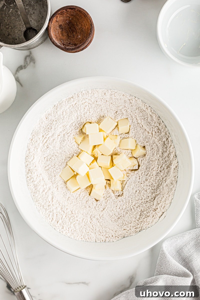 Overhead view of cold, cubed butter being cut into the flour mixture in a white mixing bowl with a pastry blender, showing the ideal 'pea-sized' consistency for flaky pie crust.