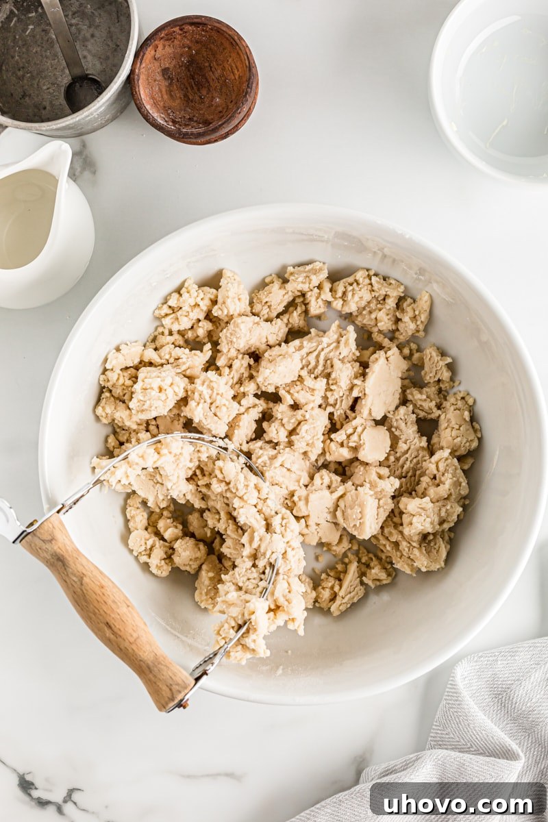 Overhead view of pie crust dough in a white mixing bowl, showing the shaggy, clumpy texture after cold water has been added and gently mixed, indicating it's almost ready for chilling.