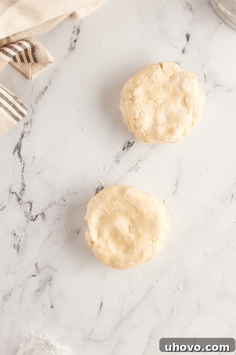 Overhead view of two portions of pie crust dough flattened into uniform discs and wrapped in plastic, ready for chilling in the refrigerator.