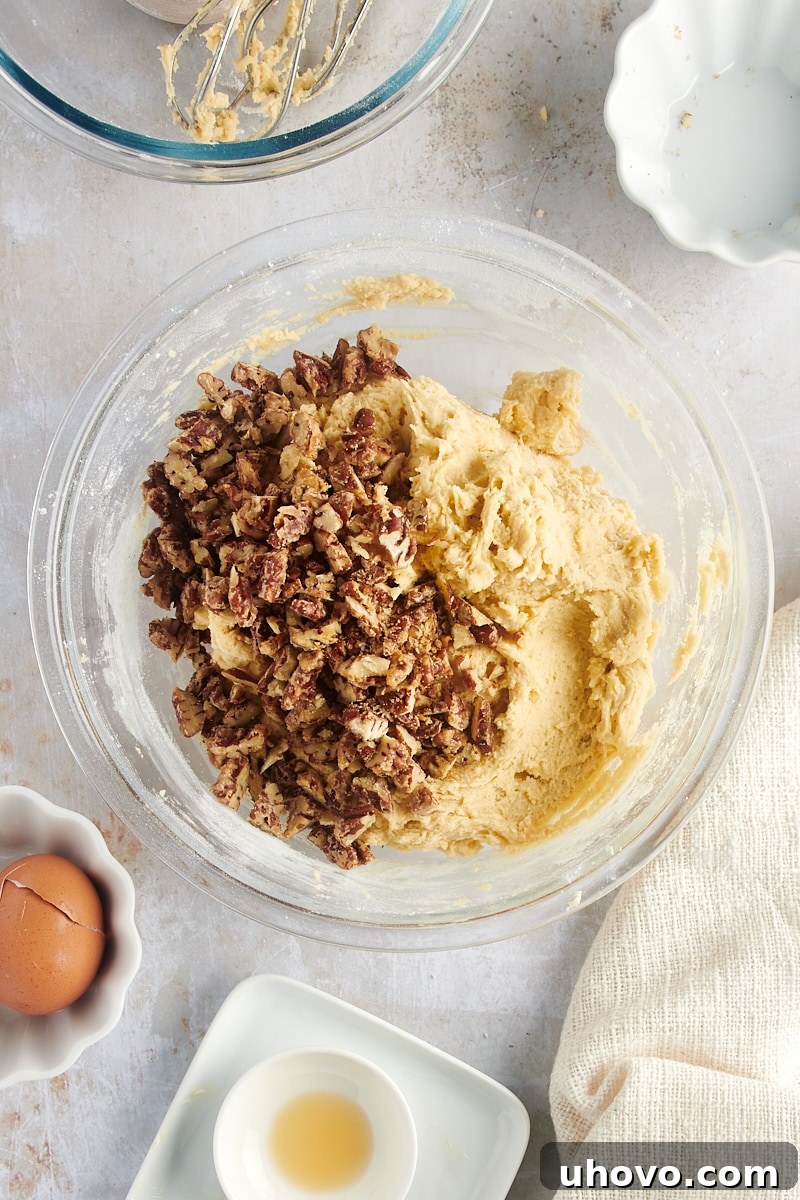 Overhead view of chopped glazed pecans being added to the cookie dough in a glass mixing bowl.