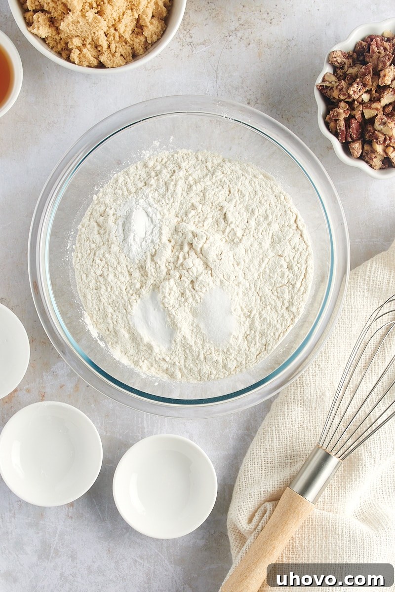Overhead view of flour, baking powder, baking soda, and salt in a clear glass mixing bowl, before combining.