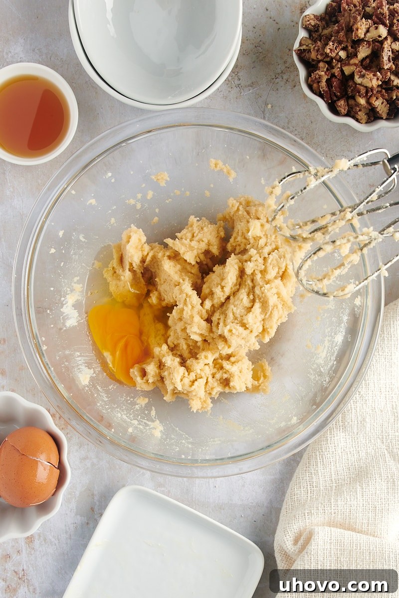 Overhead view of an egg being added to the creamy cookie dough mixture in a glass mixing bowl, ensuring proper emulsification.