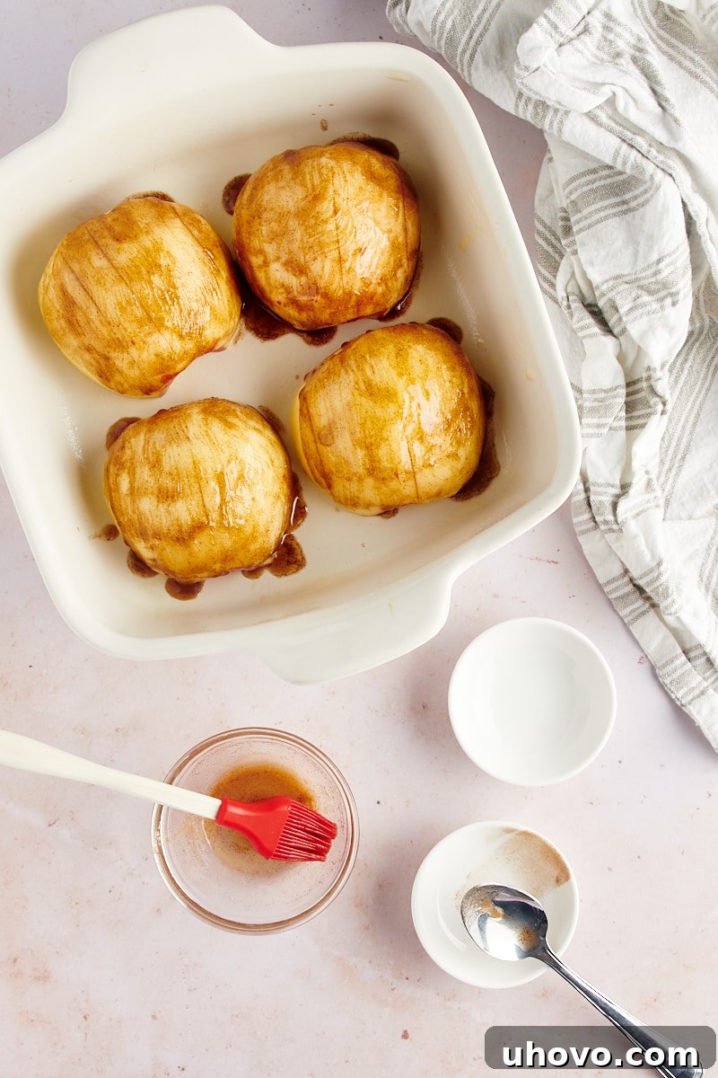 Overhead view of a white baking dish containing four apple halves, each meticulously hasselback-sliced and brushed with a glistening mixture of melted butter, brown sugar, and cinnamon