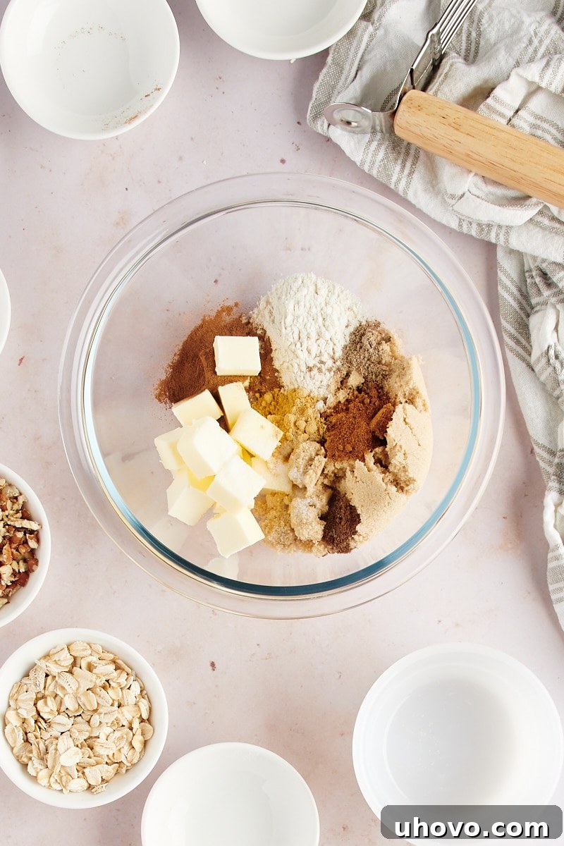 Overhead view of cold butter cubes, all-purpose flour, and a selection of ground spices (cinnamon, ginger, nutmeg, cardamom, allspice) in a clear glass mixing bowl, ready to be combined for the topping