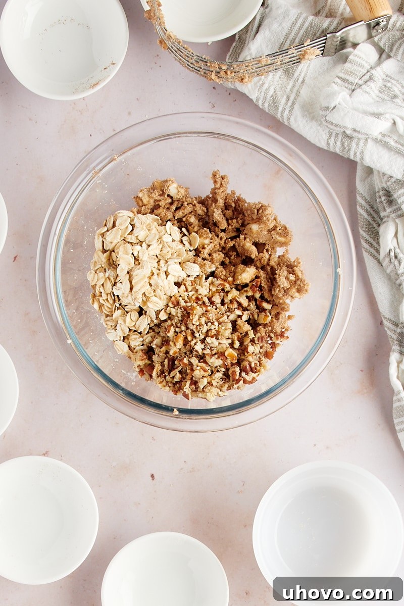 Overhead view of chopped nuts and rolled oats added to the crumb topping mixture in a glass bowl, ready for final mixing
