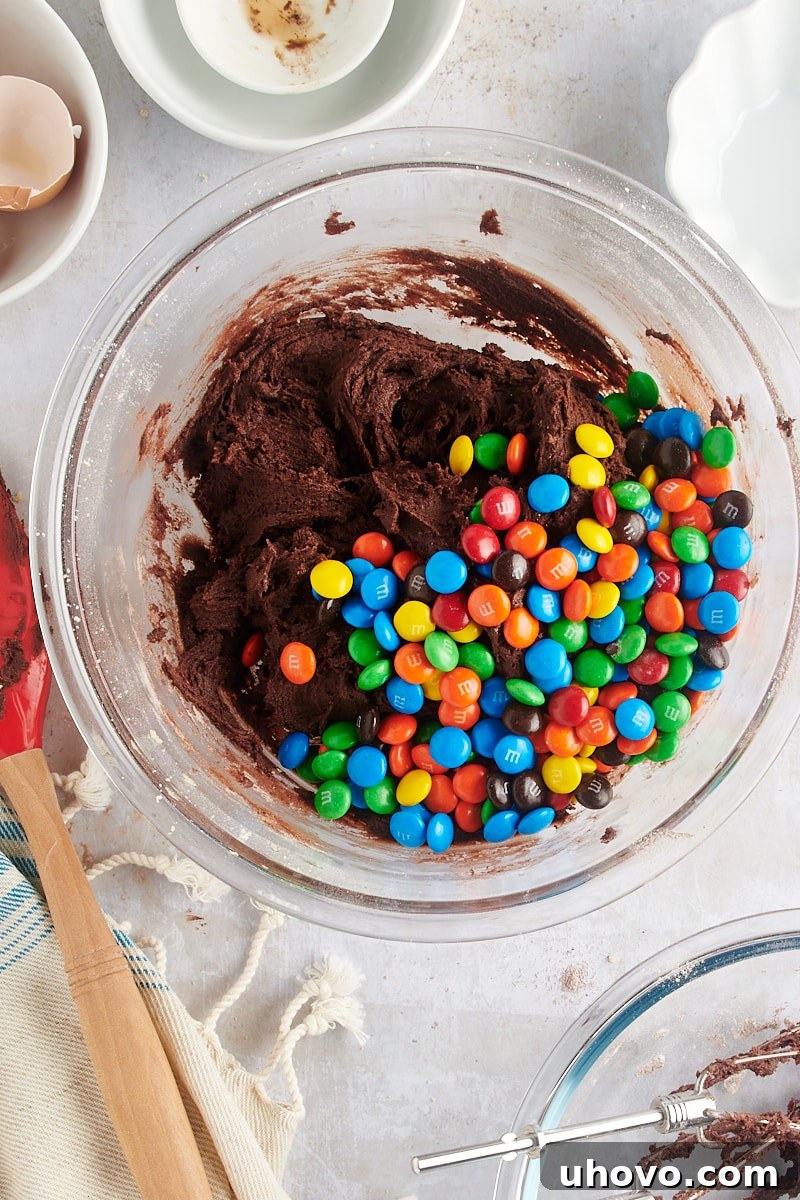 Overhead view of colorful M&Ms being gently folded into chocolate cookie dough in a glass mixing bowl.