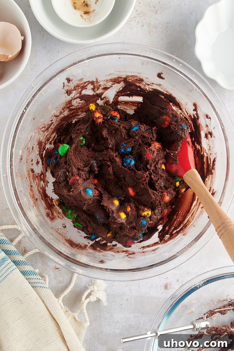 Overhead view of the fully mixed Chocolate M&M Cookie dough, rich brown with colorful candies, in a glass mixing bowl.