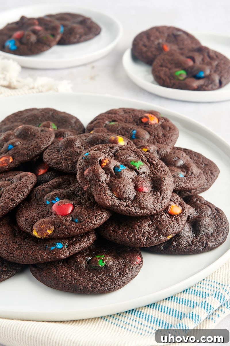 A large white plate overflowing with Chocolate M&M Cookies, with smaller plates of cookies blurred in the background, creating an inviting display.