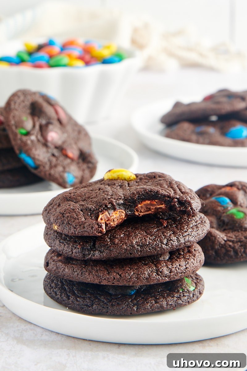 A stack of four Chocolate M&M Cookies on a small white plate, with a bite taken out of the top cookie, revealing its soft, fudgy interior and colorful candies.