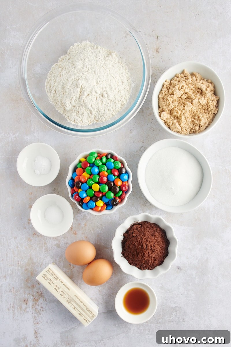 Overhead view of perfectly measured ingredients for Chocolate M&M Cookies laid out on a white surface, including flour, cocoa powder, butter, sugars, eggs, vanilla, and a bowl of M&Ms.