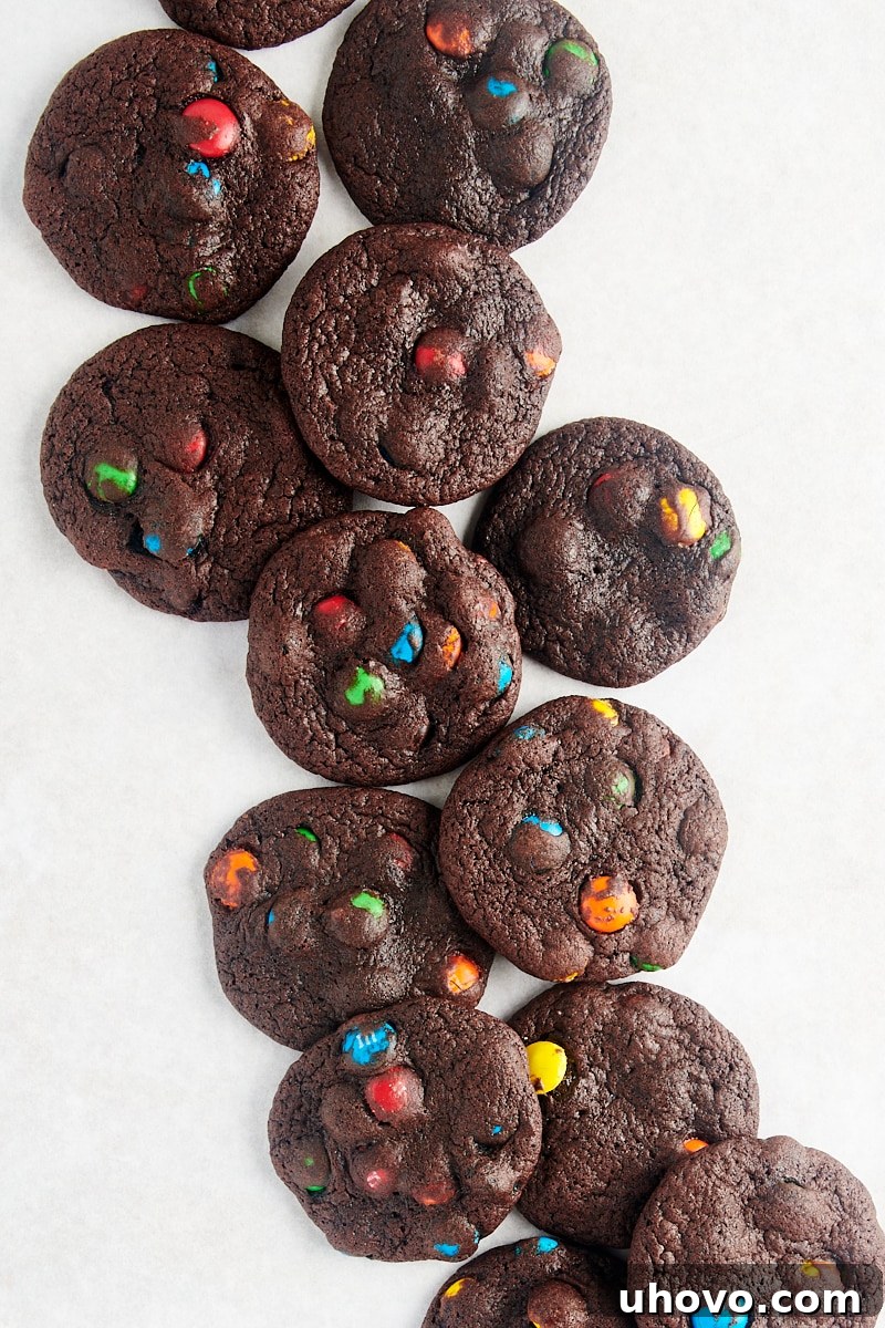 Overhead shot of freshly baked Chocolate M&M Cookies scattered invitingly across a pristine white surface, showcasing their rich color and vibrant candy toppings.