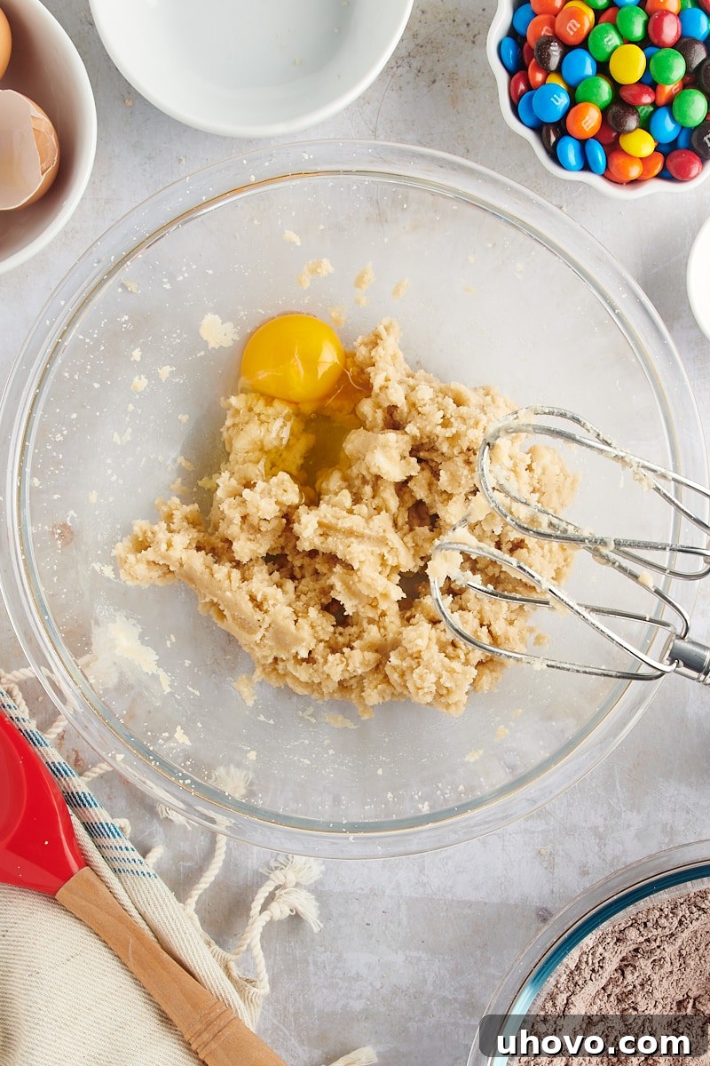 Overhead view of a single egg being added to the creamed butter and sugar mixture in a glass mixing bowl.
