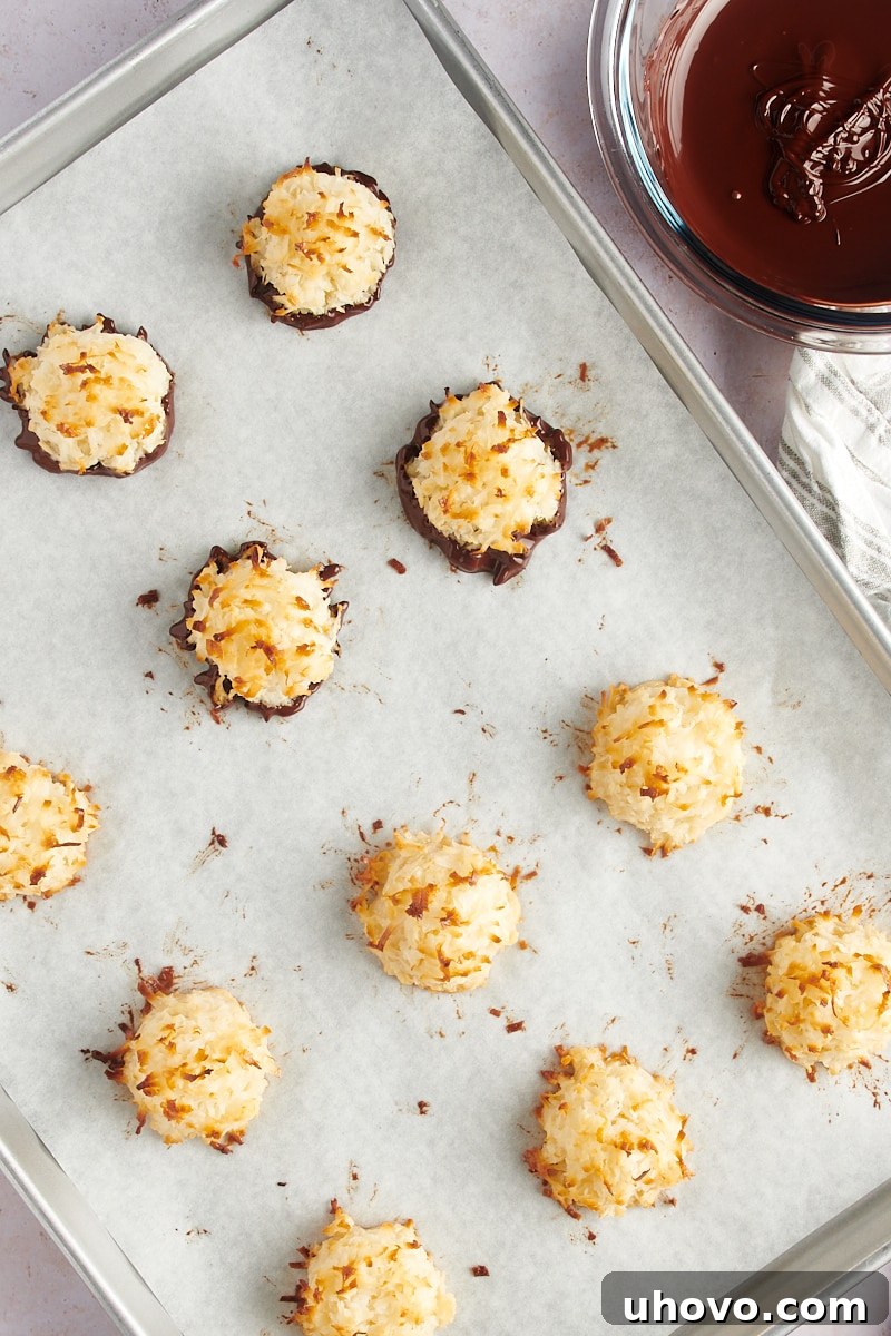 An inviting overhead view of a batch of Coconut Macaroons resting on parchment paper, with several already dipped in lustrous melted dark chocolate, ready for setting.