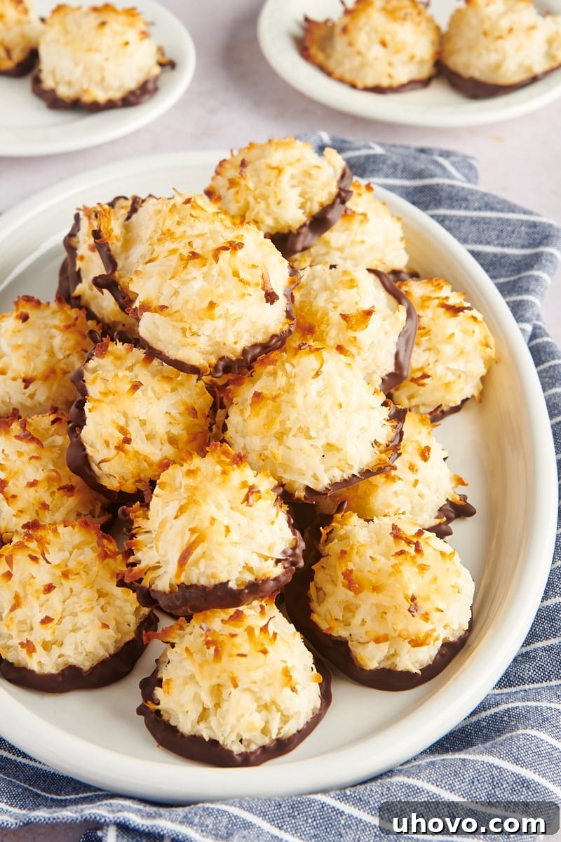 A generous pile of golden-brown Coconut Macaroons, some dipped in chocolate, beautifully presented on an oval white plate, showcasing a delicious dessert.