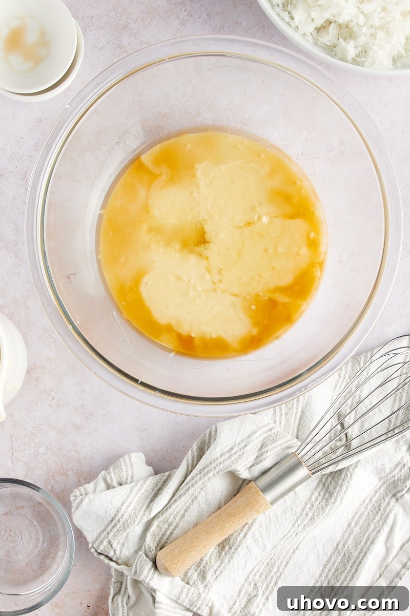 An overhead view of sweetened condensed milk, separated egg whites, vanilla and almond extracts, and a pinch of salt waiting in a clear glass mixing bowl, ready to be combined.