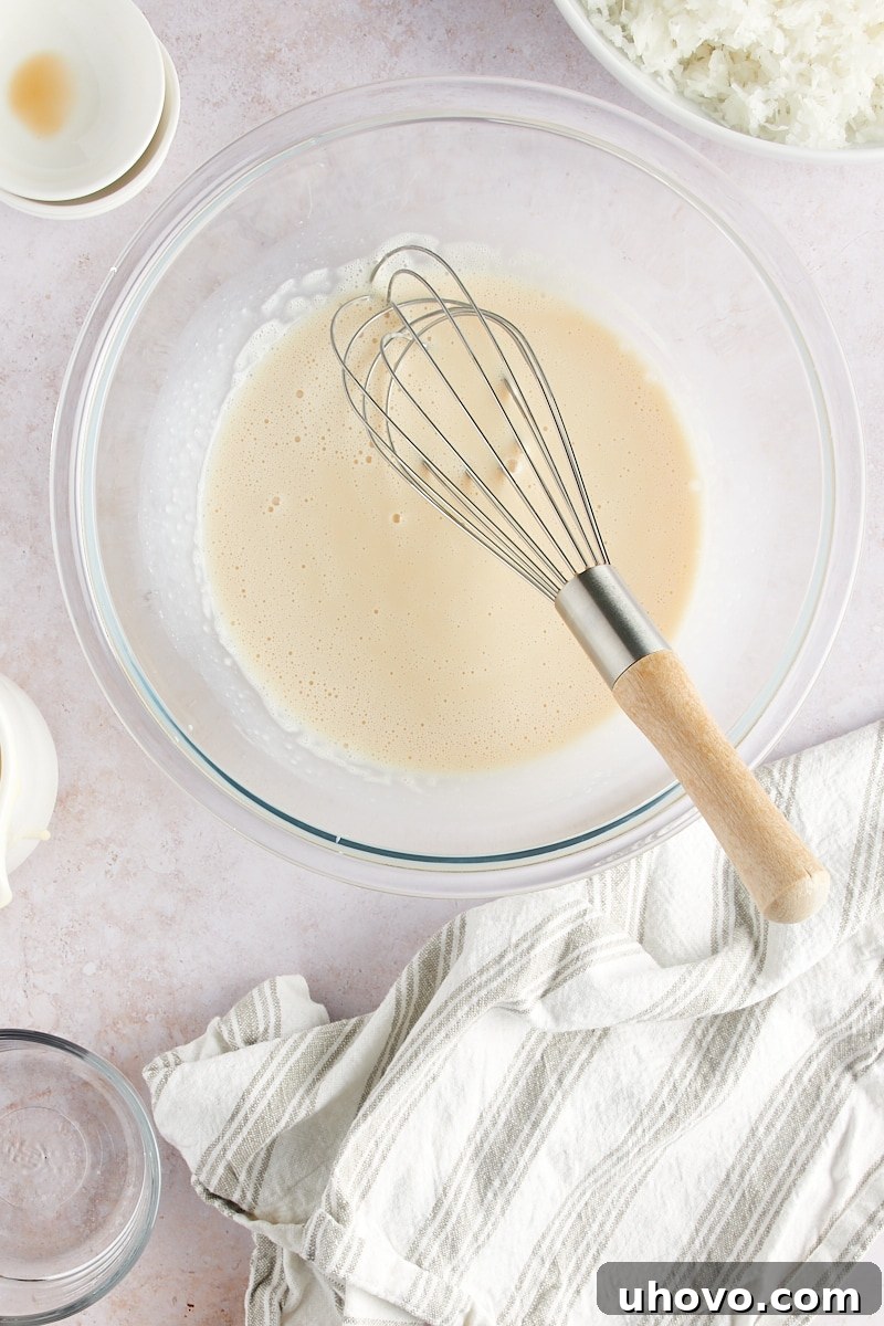 An overhead shot of the wet ingredients for Coconut Macaroons – condensed milk, egg whites, and extracts – expertly whisked together into a smooth, slightly frothy mixture in a glass bowl.