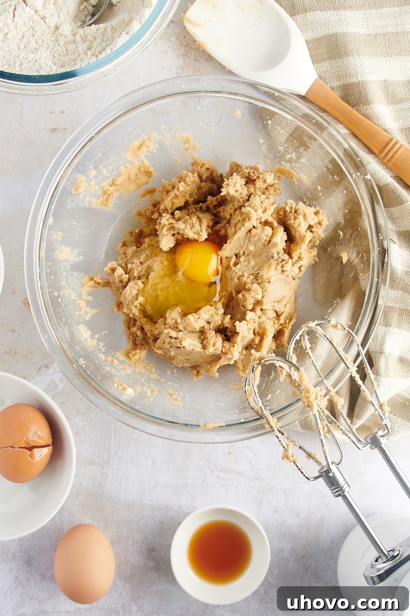 Overhead view of a cracked egg added to the creamed mixture of butter and sugars in a glass mixing bowl, before being fully incorporated.