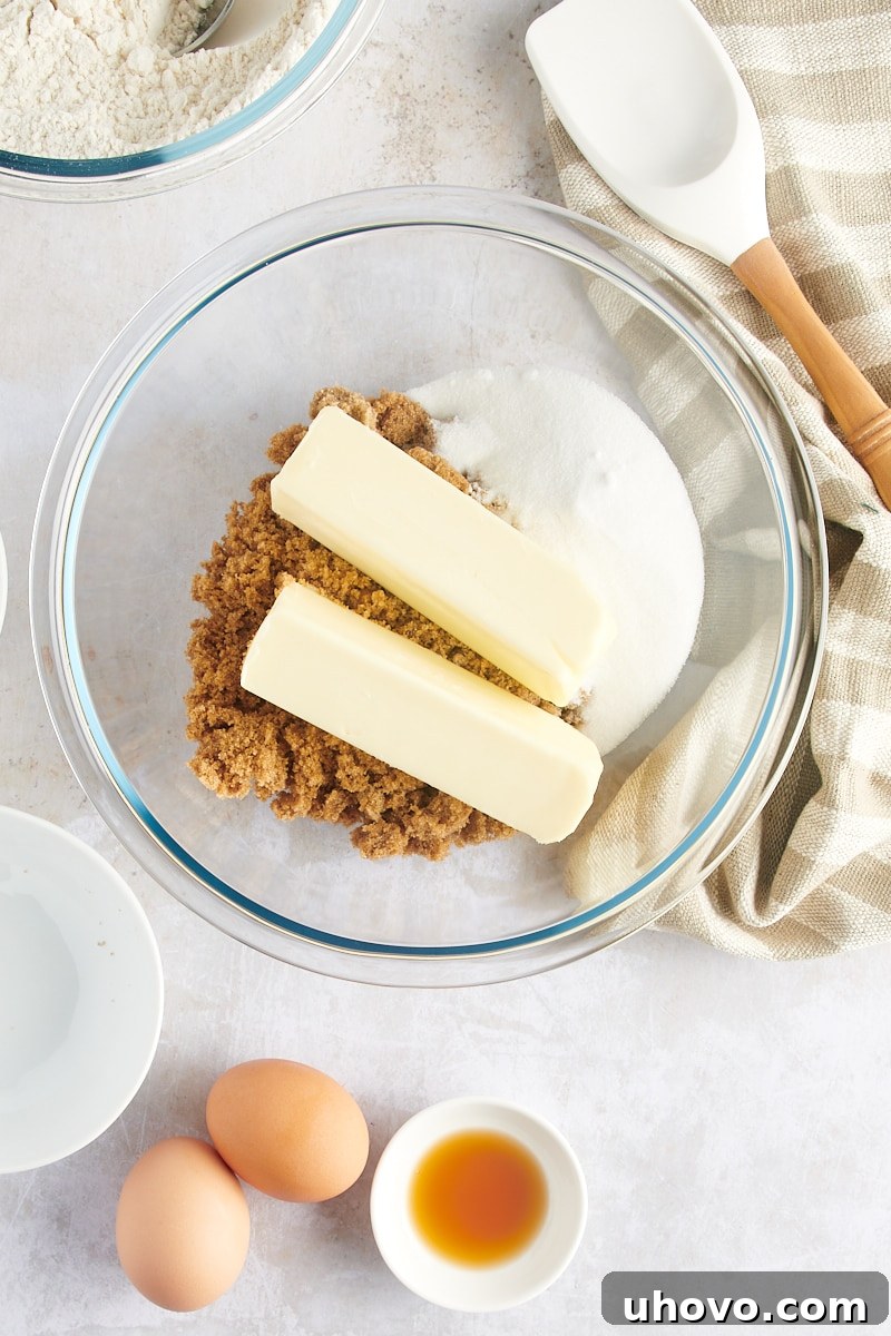 Overhead view of softened unsalted butter, dark brown sugar, and granulated sugar in a large glass mixing bowl, ready for creaming.