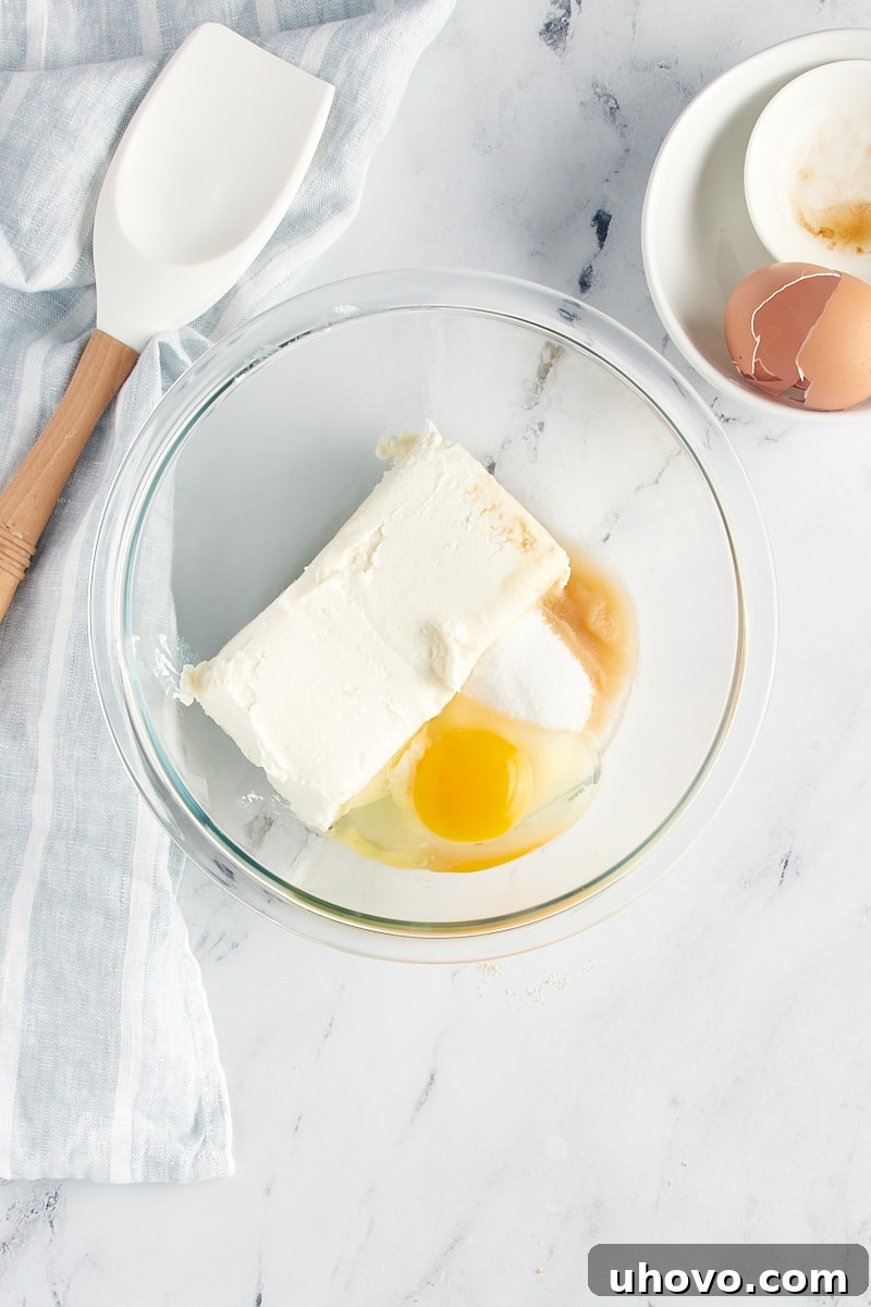 All the cheesecake filling ingredients – cream cheese, sugar, egg, and vanilla – combined in a glass mixing bowl, ready to be beaten.