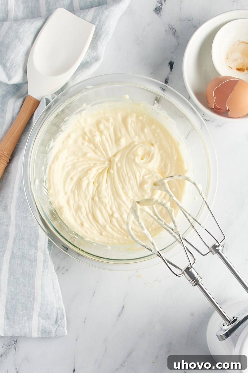 A handheld electric mixer actively beating the cheesecake filling in a glass bowl until it reaches a perfectly smooth consistency.